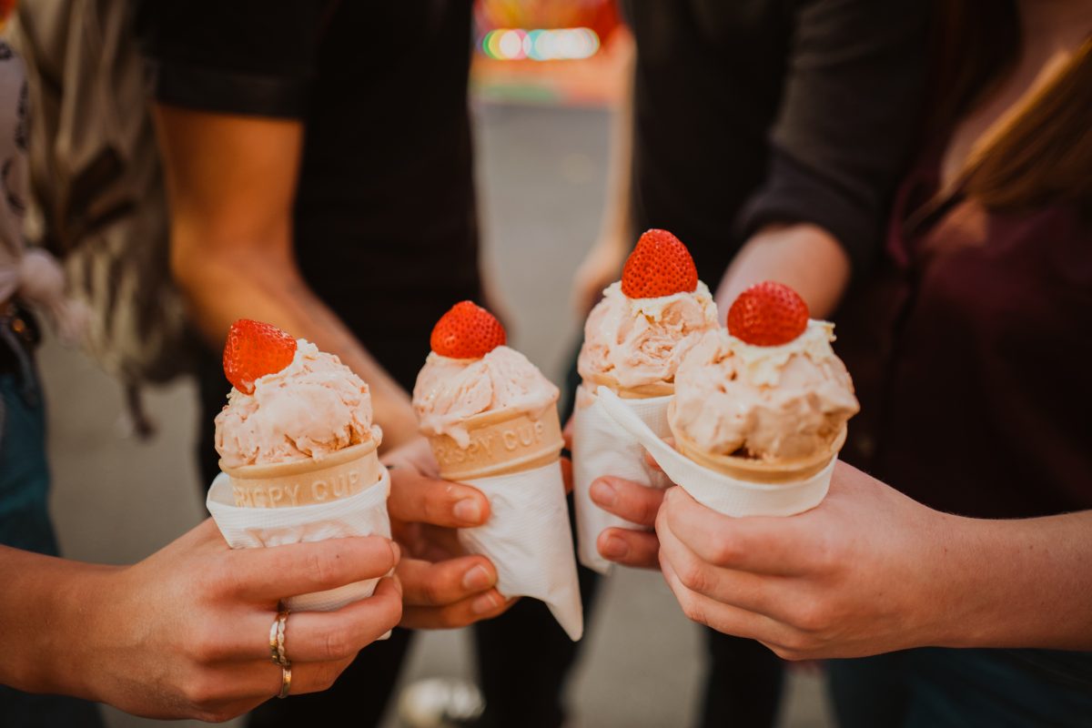 A close up of four hands, each holding a strawberry ice cream cone with a fresh strawberry on top.