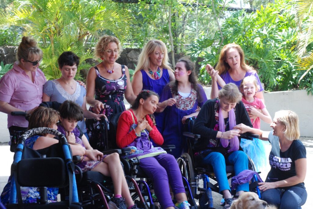 Image is of a group of people dressed in purple. Six people are standing in the back row. The front row has women in wheelchairs and two people crouching on either side of them. The head of a dog is in the bottom of the photo.